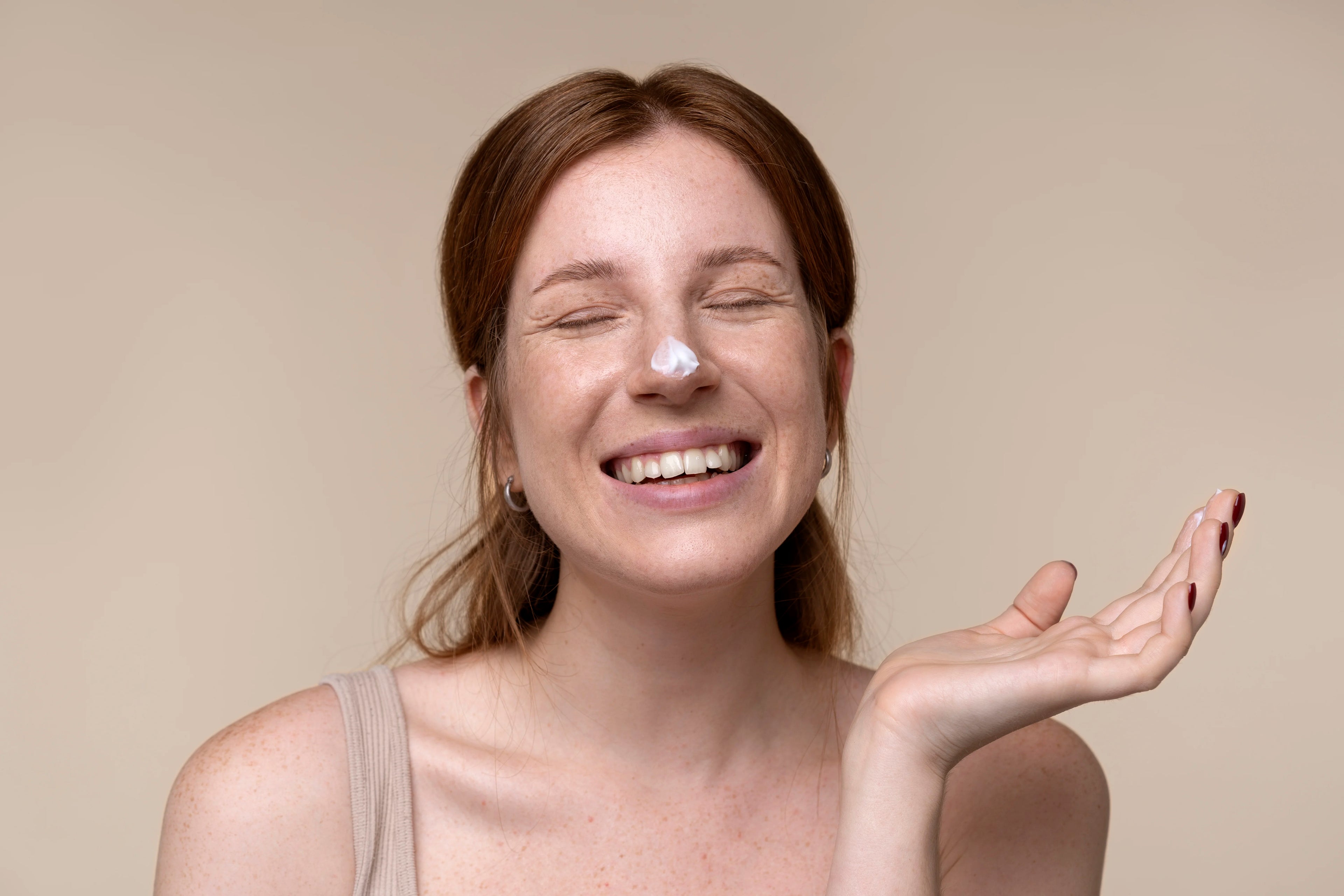 Woman with a smile on her face, showing cream on her hand against a beige background.